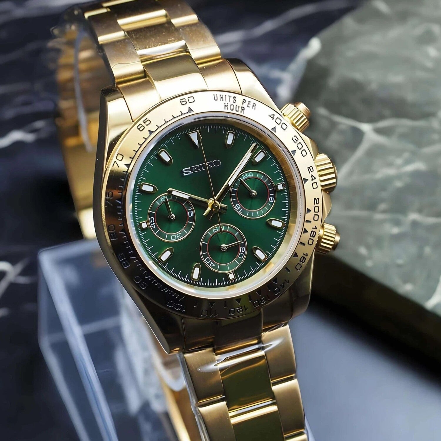 Angled view of the Seiko Mod Daytona in polished gold with rich green dial, three chronograph subdials, and matching gold bracelet displayed on a marble surface.