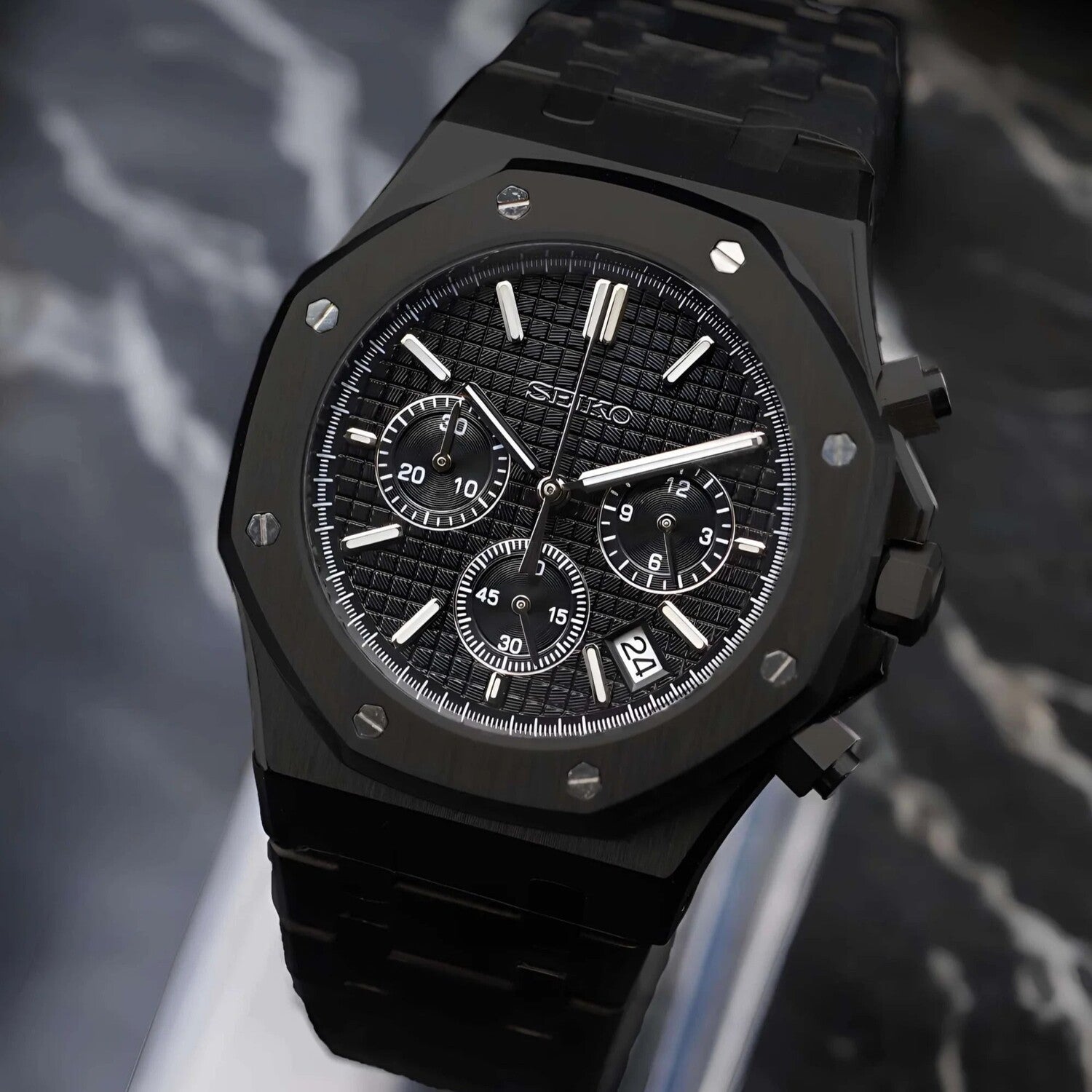 Angled view of the Seiko Mod Royal Oak VK63 chronograph in triple black with a textured dial, three subdials, matte stainless steel bracelet, chronograph pushers, and a date display positioned between 4 and 5 o’clock