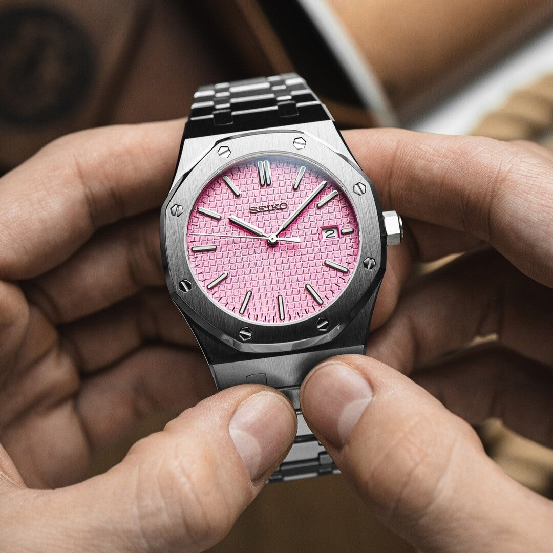 Top view of a Seiko Mod Royal Oak with brushed stainless steel case, pink textured dial, applied silver hour markers, date window at 3 o’clock, and integrated steel bracelet held in both hands