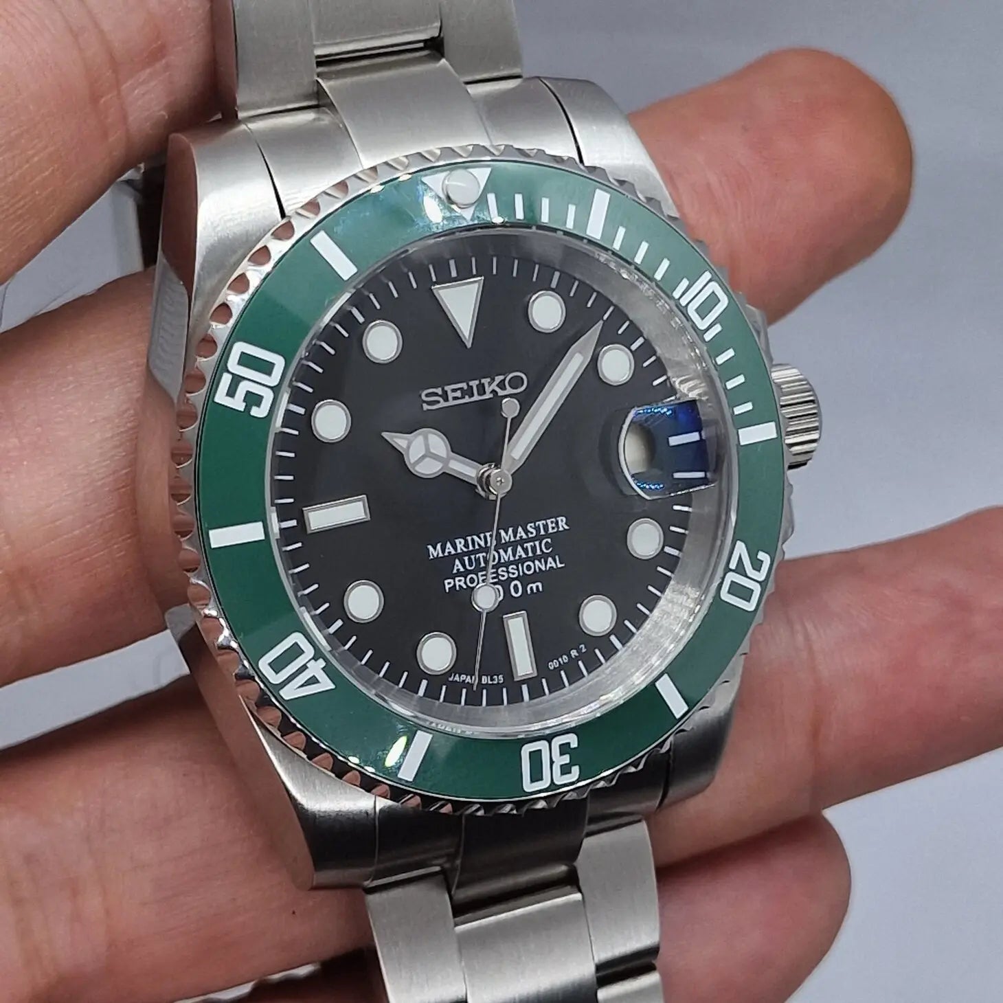 Nahaufnahme der Seiko Mod Submariner Kermit, gehalten in der Hand. Die Taucheruhr zeigt ihre gruene Luenette, das schwarze Zifferblatt mit Datumslupe und das gebuerstete Edelstahlarmband in leichter Schraegansicht.