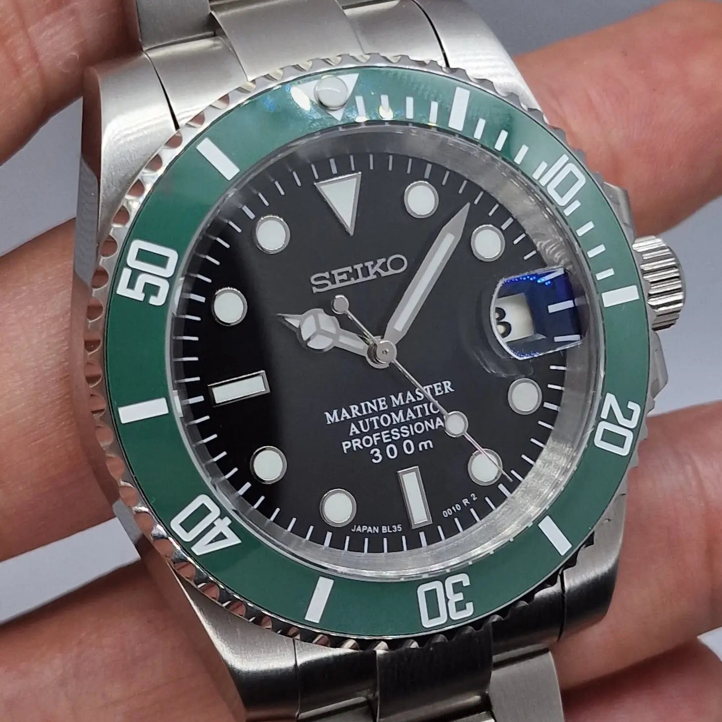 Detailaufnahme der Seiko Mod Submariner Kermit aus linker Perspektive. Das Bild zeigt die gruene Luenette, das schwarze Zifferblatt mit Datumslupe und die praezise Verarbeitung der automatischen Taucheruhr aus Edelstahl.