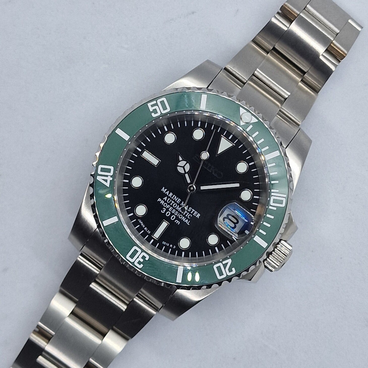 Obenansicht der Seiko Mod Submariner Kermit mit schwarzem Zifferblatt, gruener Luenette und Edelstahlarmband. Das Bild zeigt die automatische Taucheruhr mit Datumslupe und 300m Wasserdichtigkeit auf hellem Hintergrund.