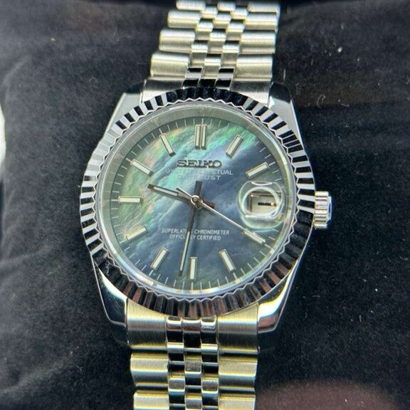 Seiko Mod Datejust Cosmos mit schimmerndem Perlmutt-Zifferblatt in Blau-Grün-Tönen, gerillter Edelstahl-Lünette, Jubilee-Armband und Datumsanzeige auf schwarzem Uhrenkissen