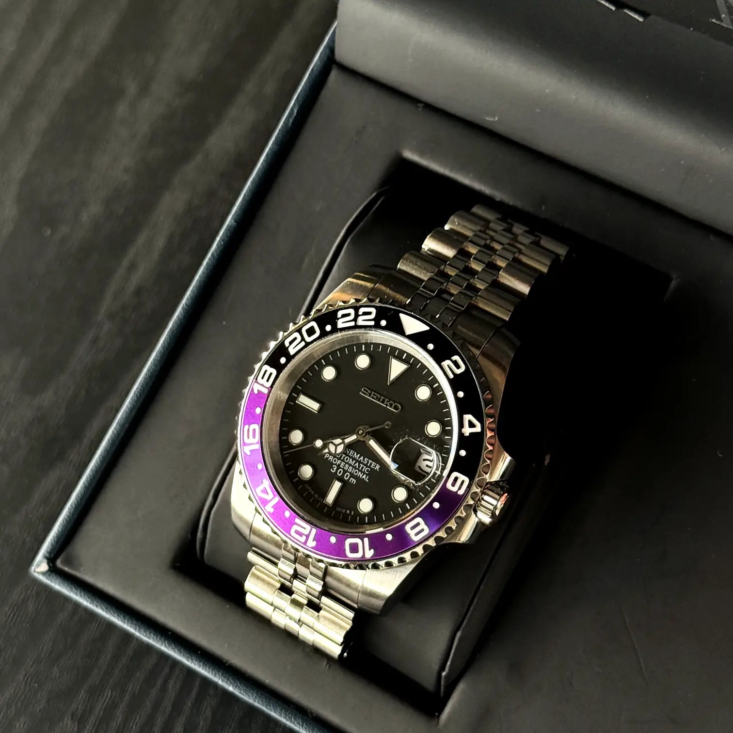 Seiko Mod GMT Joker in offener Box. Uhr mit schwarzem Zifferblatt, violett-schwarzer GMT-Lünette und Jubilee-Edelstahlarmband.