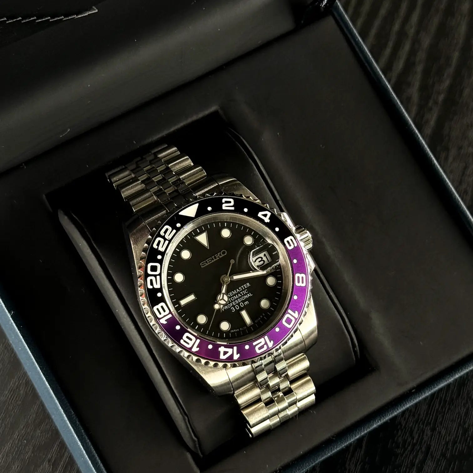 Seiko Mod GMT Joker in offener Verpackung. Automatikuhr mit schwarzem Zifferblatt, violett-schwarzer GMT-Lünette und Jubilee-Edelstahlarmband